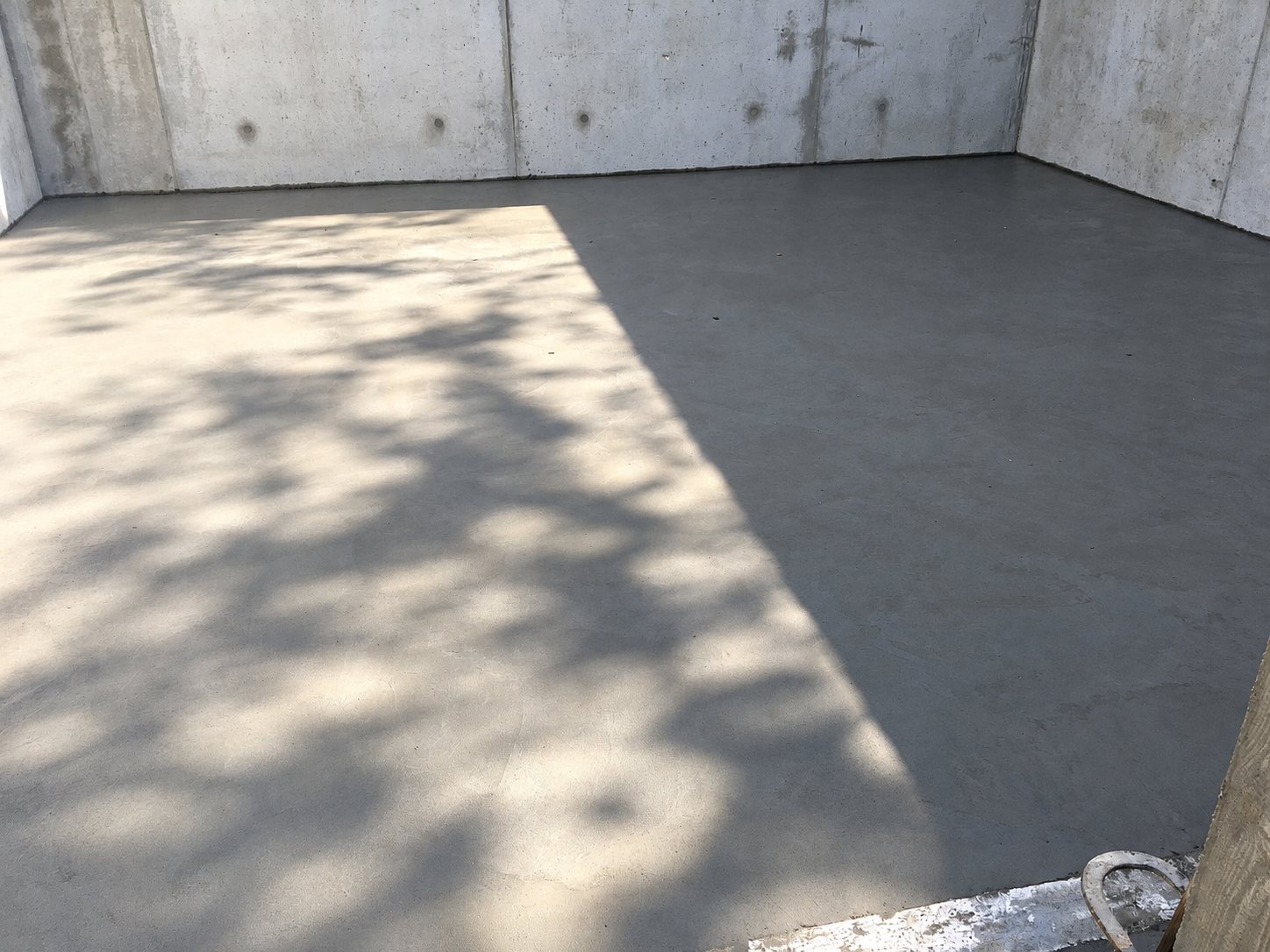Une dalle de béton lisse fraîchement coulée, partiellement éclairée par le soleil avec des ombres de feuillage, entourée de murs en béton brut.