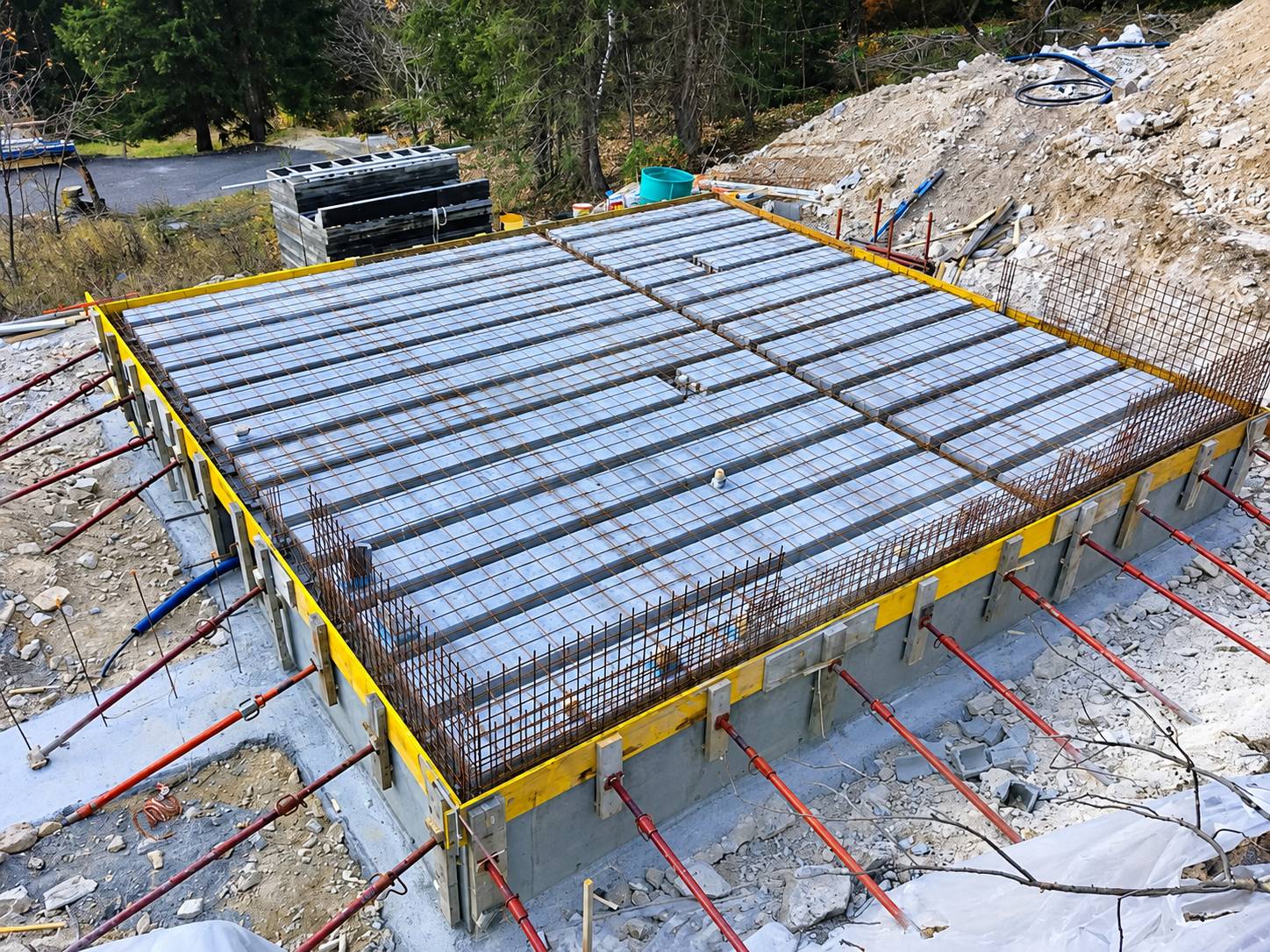 Vue en hauteur d'une dalle de plancher en cours de construction, avec treillis d'armature, banches jaunes et étais rouges, au milieu d'un terrain rocailleux en bord de forêt.