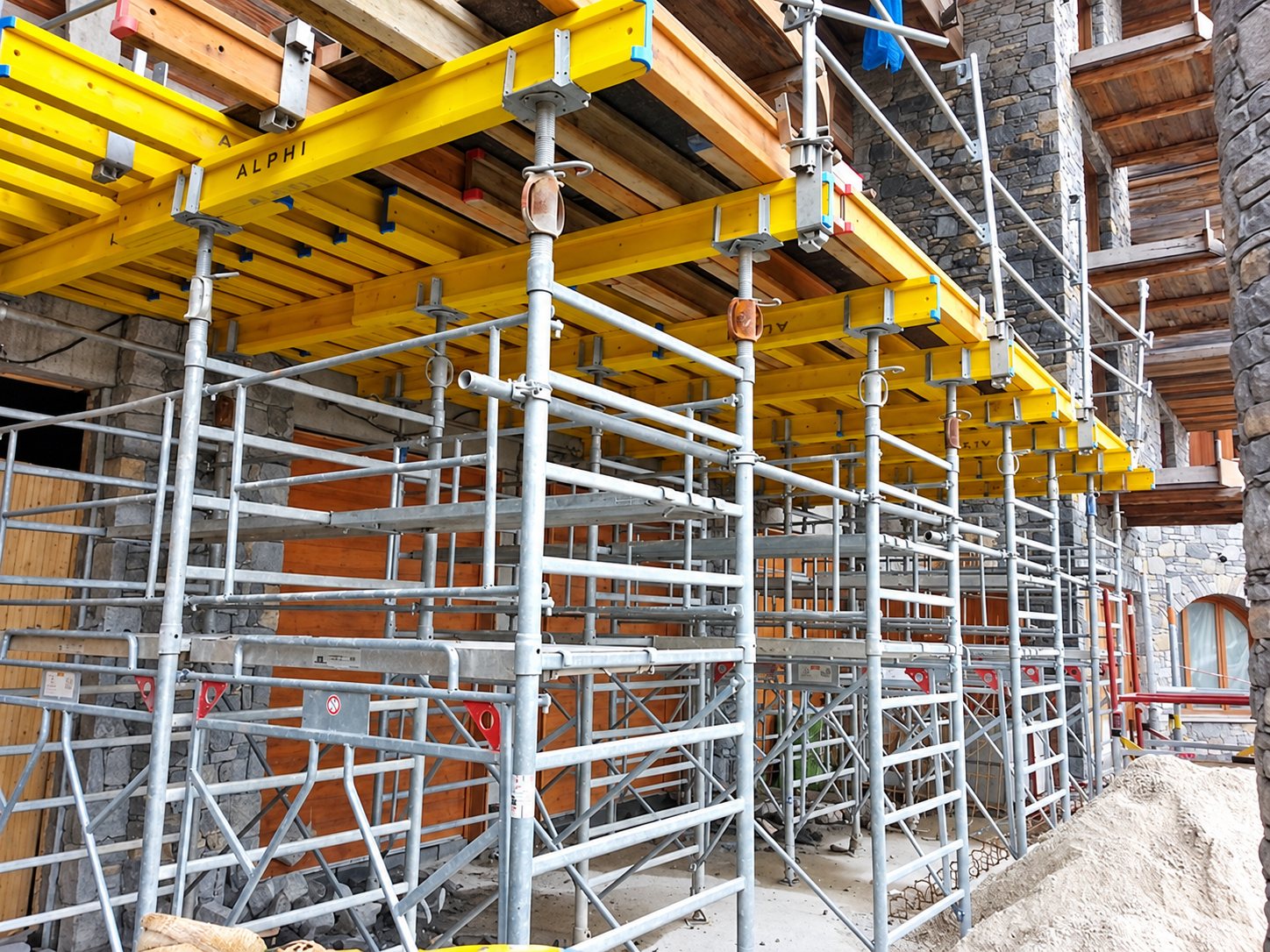 Structure d'échafaudage métallique supportant des poutres de coffrage jaunes devant un bâtiment en pierre et bois sur un chantier.