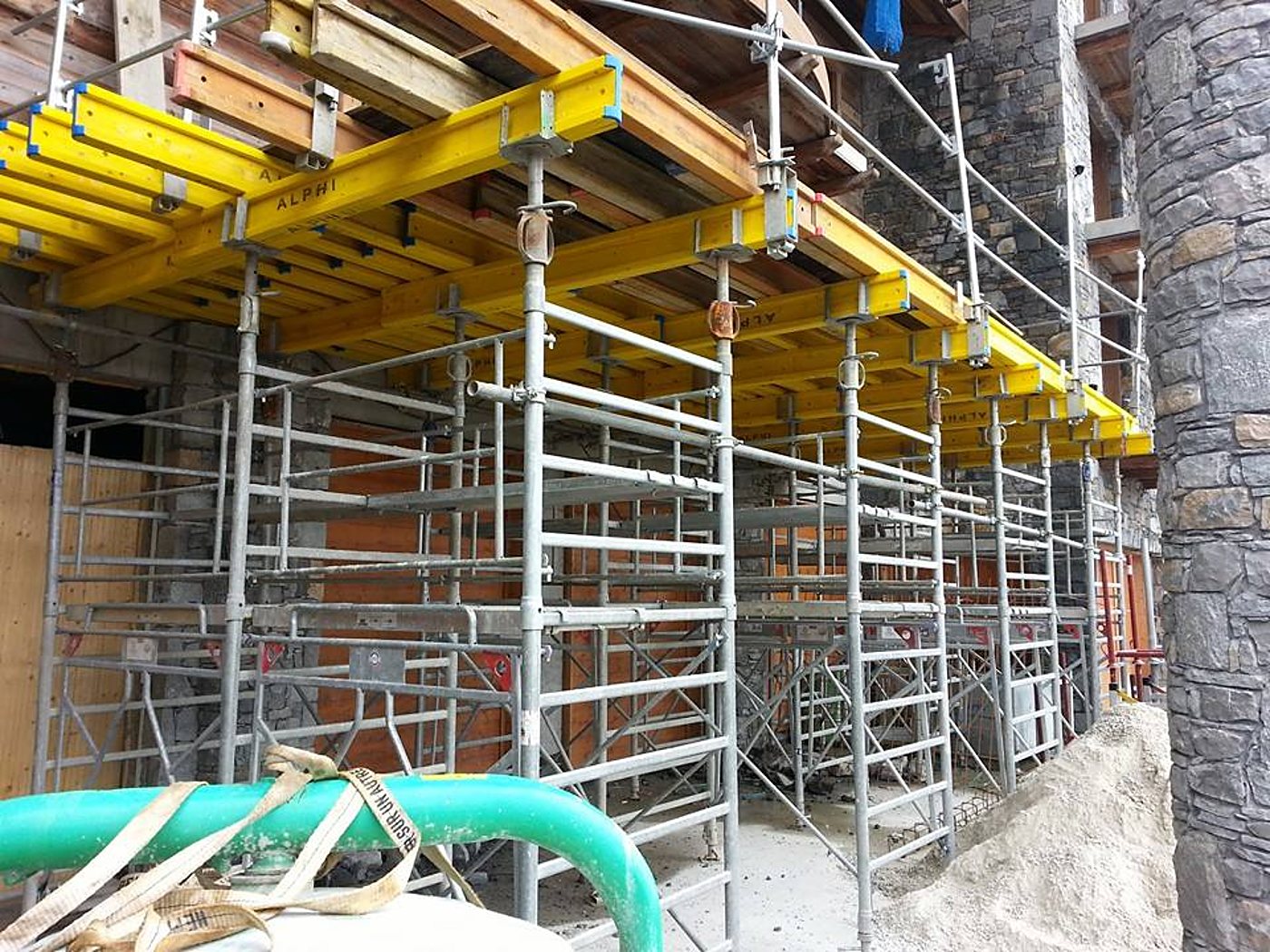 Des poutrelles jaunes de coffrage reposent sur des étais et des échafaudages métalliques sur un chantier, près d'un mur en pierre et d'un tas de sable.