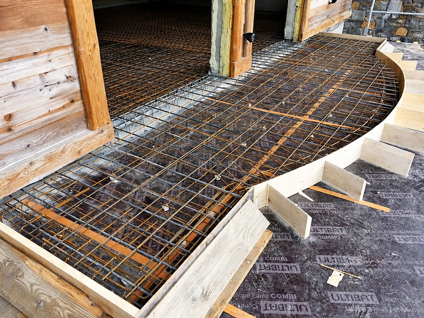 Treillis de fer à béton posé dans un coffrage en bois courbe sur une terrasse en construction, devant un mur de chalet en bois.
