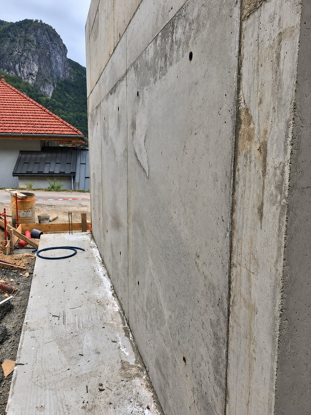 Un grand mur en béton brut longe une dalle sur un chantier, avec une maison au toit rouge et une montagne en arrière-plan.