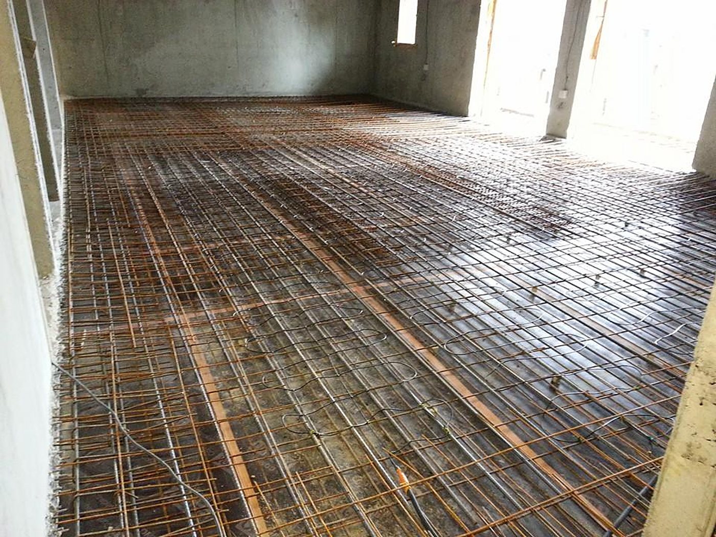 Pièce en béton brut dont le sol est couvert d'un treillis d'armature en acier sur lequel sont posés des câbles de plancher chauffant.